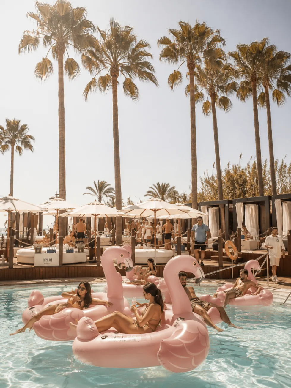 Guests floating on pink inflatables in the pool at Opium Beach Marbella beneath palms and cabanas