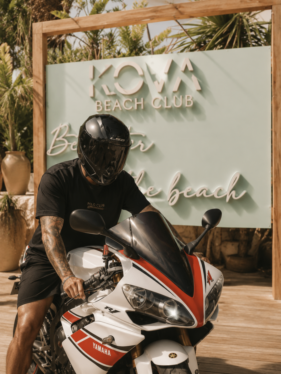 Arrival sign at Kova Beach Club with a motorbike parked in front and tropical planting around the entrance